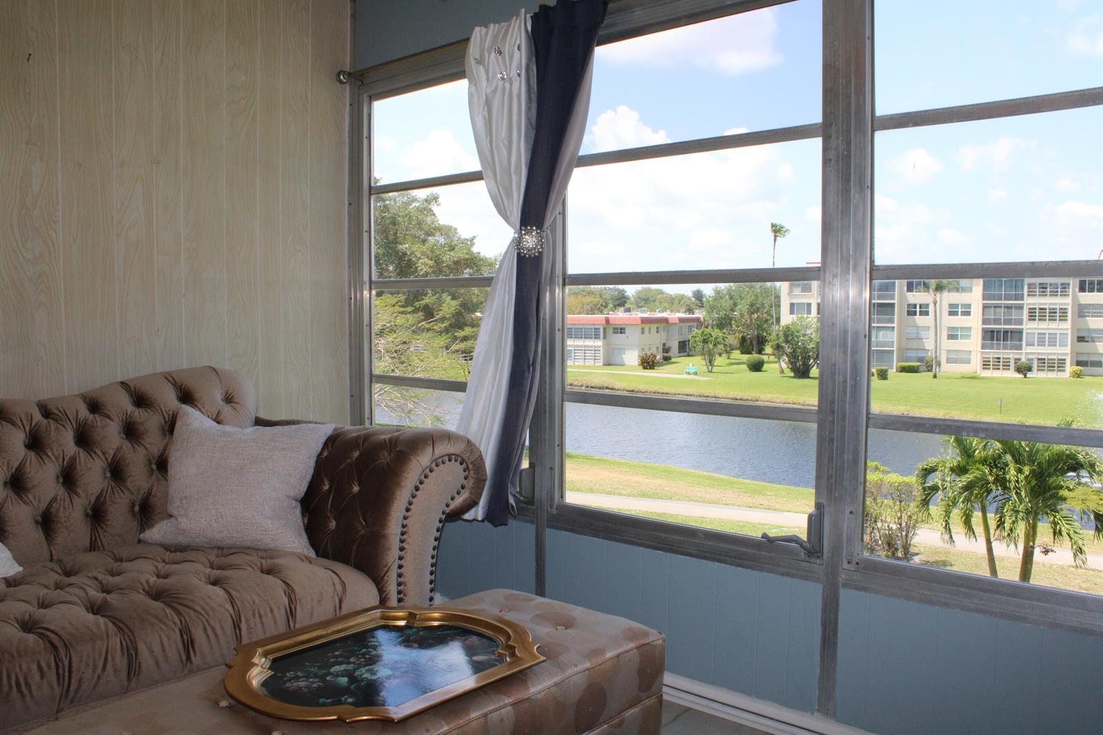 A sunroom features a tufted brown sofa, an ottoman, and large windows overlooking a canal — 14671 Bonaire Boulevard # 305, Delray Beach FL 33446, DELRAY BEACH, FL
