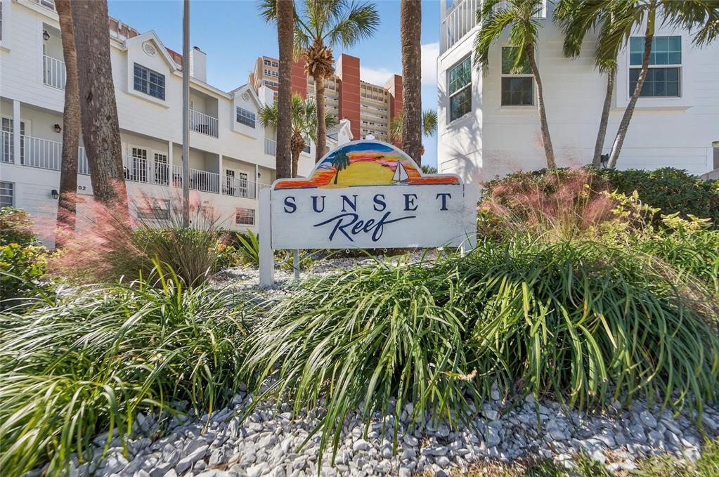 The exterior of the Sunset Reef complex features a white sign surrounded by landscaping — 17960 GULF BLVD UNIT 108, REDINGTON SHORES, FL