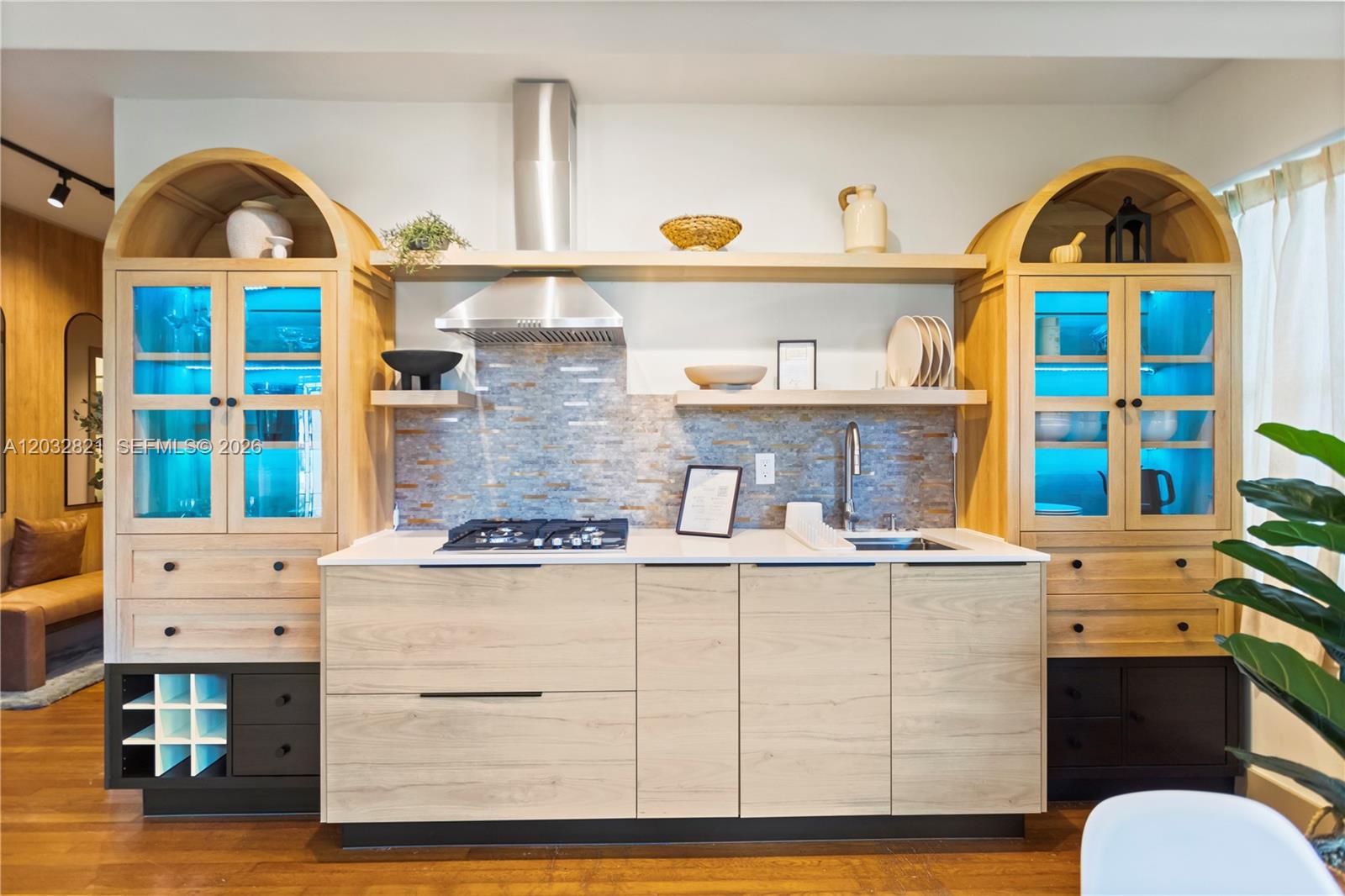 A modern kitchen features light wood cabinetry, blue-lit glass cabinets, and a stone tile backsplash — 499 NE 112th St, Miami FL 33161, MIAMI, FL