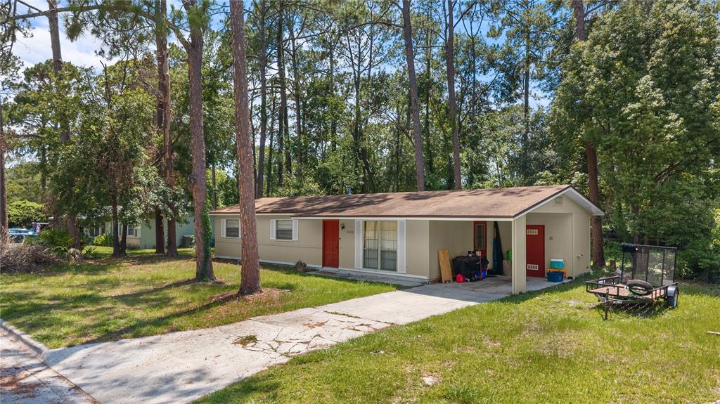 A single-story house with a tan exterior, red doors, and a carport sits among trees — 5030 NW 28TH TERRACE, GAINESVILLE, FL