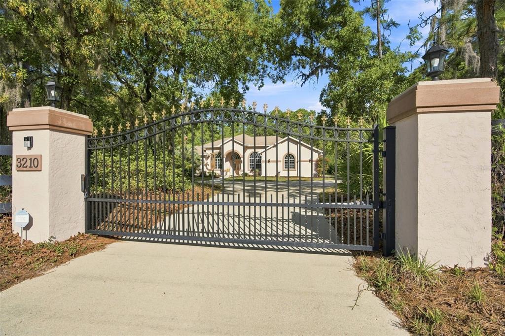 A gated driveway leads to a single-story house surrounded by mature trees and landscaping — 3210 N RENO WAY, BEVERLY HILLS, FL
