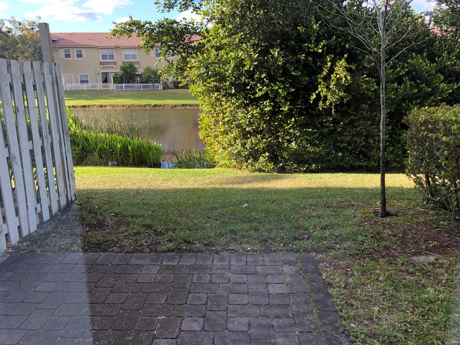 A paved patio overlooks a grassy backyard, a pond, and residential buildings in the distance — 261 Lake Monterey Circle, Boynton Beach FL 33426, BOYNTON BEACH, FL