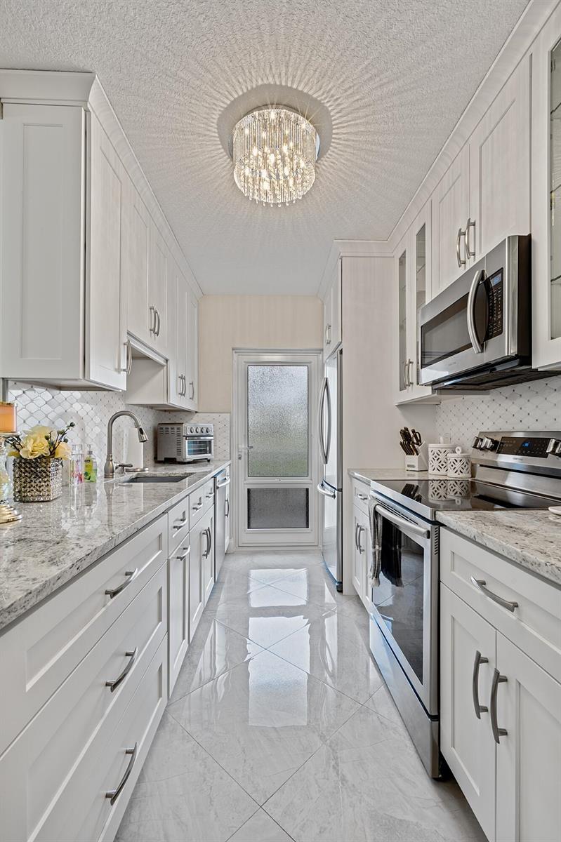 A galley kitchen features white cabinets, granite countertops, stainless steel appliances, and a crystal chandelier — 1910 S Ocean Boulevard # 204, Delray Beach FL 33483, DELRAY BEACH, FL