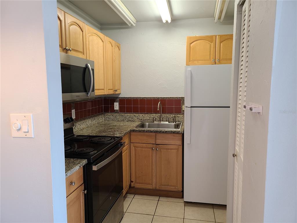 A small kitchen features light wood cabinets, granite countertops, a dark tile backsplash, and appliances — 2763 WOODGATE LANE UNIT 207, SARASOTA, FL