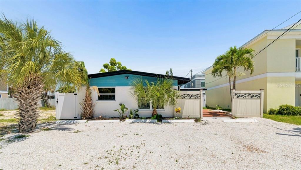 A single-story house with a white brick exterior and teal trim sits behind a gravel lot — 14906 N BAYSHORE DRIVE, MADEIRA BEACH, FL