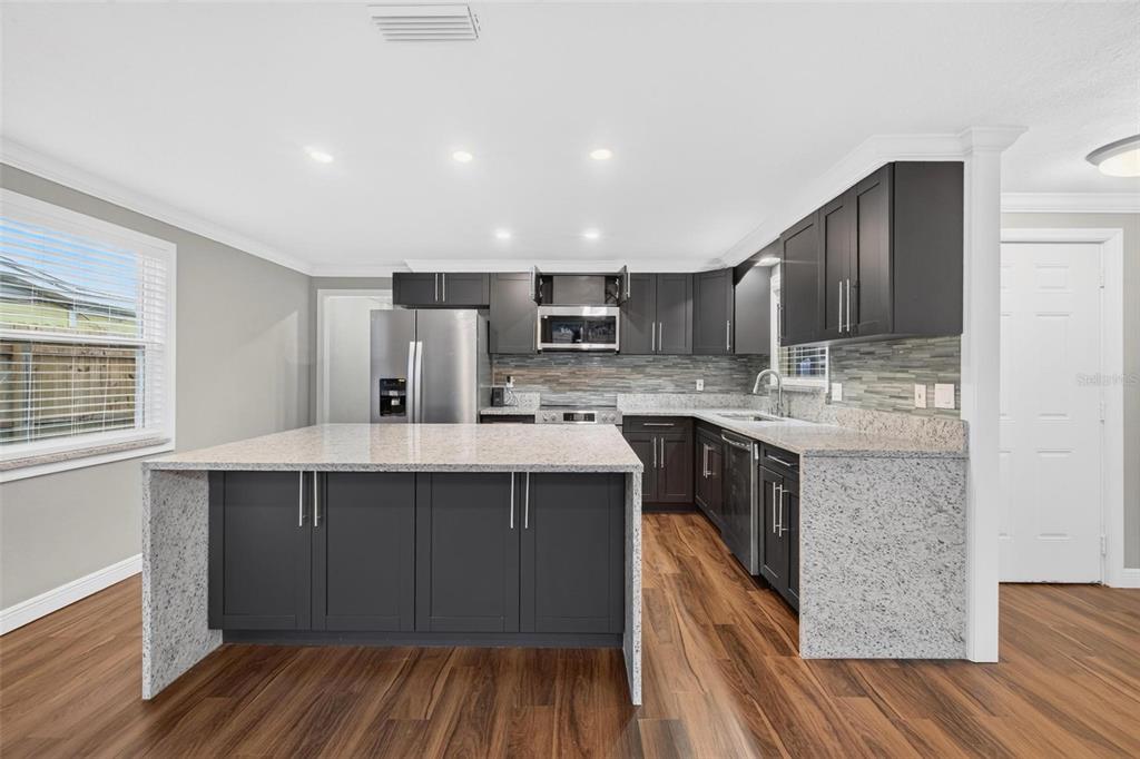 A modern kitchen features dark gray cabinets, a large granite island, and wood-look flooring — 8700 58TH LANE N, PINELLAS PARK, FL