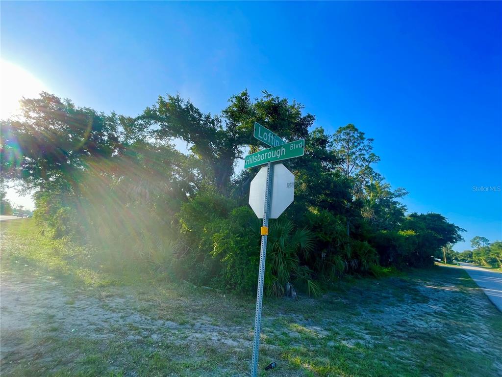 A street sign at the intersection of Loftin and Hillsborough stands before a wooded lot — 19129 HILLSBOROUGH BOULEVARD, PORT CHARLOTTE, FL
