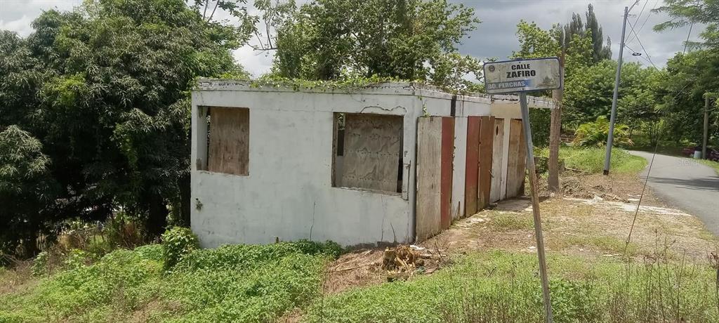 A small, weathered, white concrete structure with boarded-up windows stands near a street sign — Carr. 435 COMUNIDAD PERCHAS II, CALLE ZAFIRO, SAN SEBASTIAN, PR