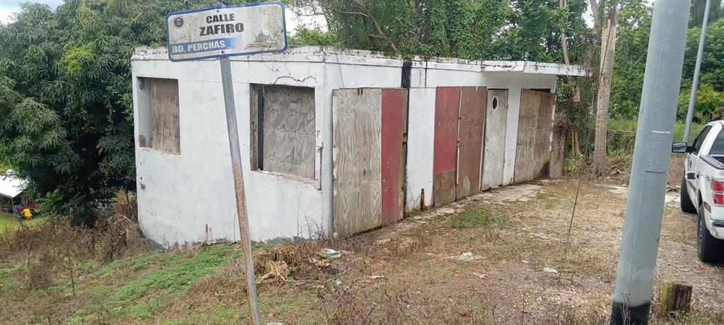 A small, weathered, white concrete building with boarded-up windows and doors sits near a street sign — Carr. 435 COMUNIDAD PERCHAS II, CALLE ZAFIRO, SAN SEBASTIAN, PR