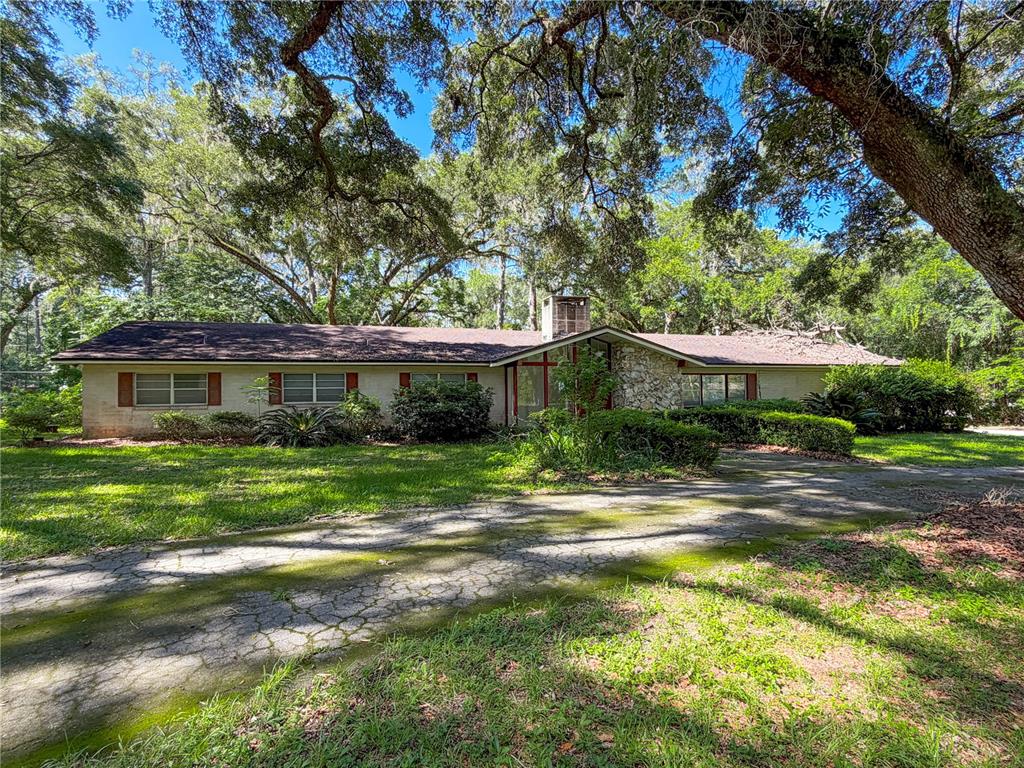 A single-story ranch-style house with a stone chimney sits behind a mossy, cracked driveway — 11131 NW 11TH AVENUE, GAINESVILLE, FL
