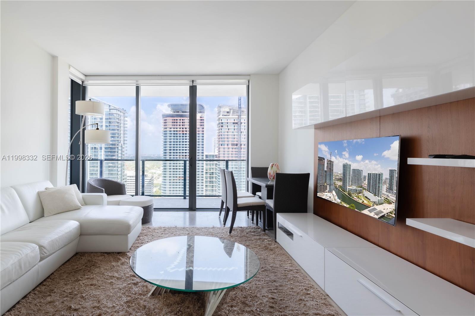 A modern living room features a white sectional sofa, glass coffee table, and city views — 68 SE 6th St # 3006, Miami FL 33131, MIAMI, FL