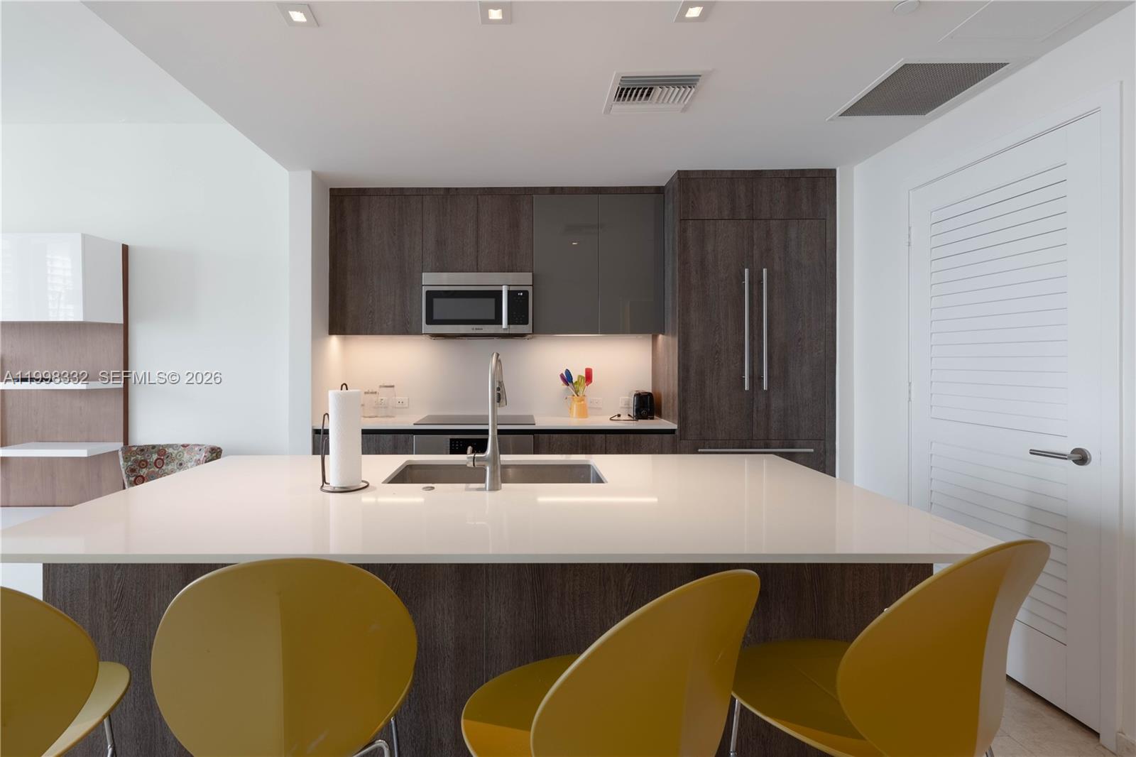 A modern kitchen features a white island, dark wood cabinetry, and yellow bar stools — 68 SE 6th St # 3006, Miami FL 33131, MIAMI, FL