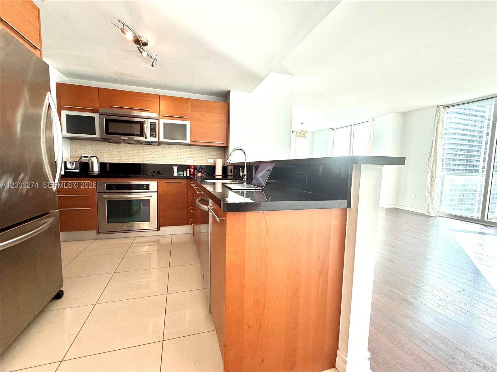 A kitchen with wood cabinets, black countertops, and stainless steel appliances opens to a living area — 244 Biscayne Blvd # 1709, Miami FL 33132, MIAMI, FL