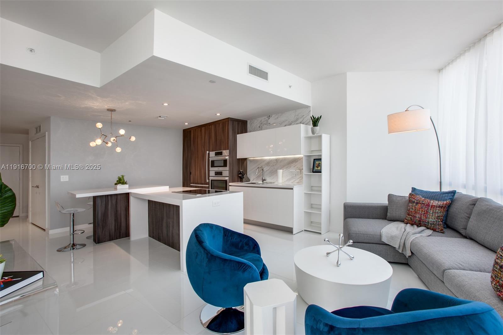 A modern open-concept living room and kitchen featuring white floors, blue chairs, and wood cabinetry — 851 NE 1ST Ave # 1810, Miami FL 33132, MIAMI, FL