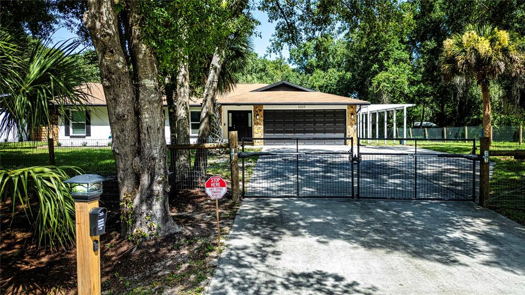 A single-story house with a black garage door behind a gated driveway and trees — 3309 YELLOWKNIFE CIRCLE, WIMAUMA, FL