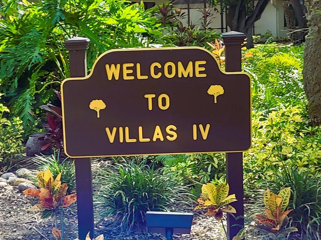 A brown sign reading Welcome to Villas IV stands in a lush, landscaped garden area — 6402 MEADOWLARK LN, BRADENTON, FL