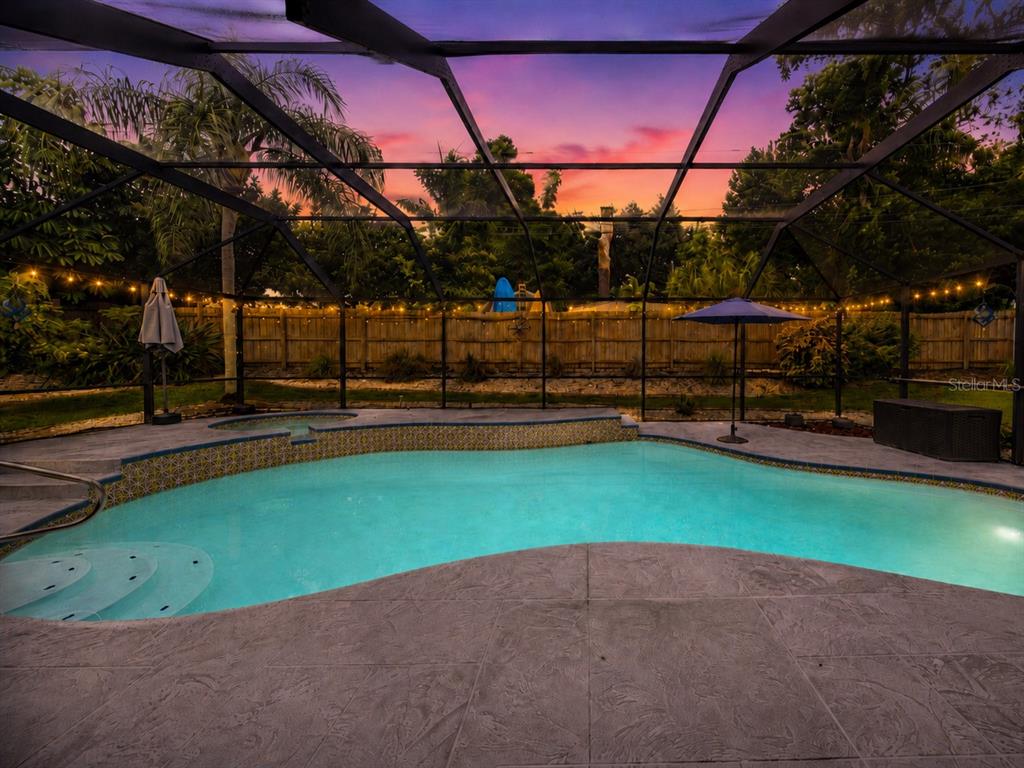 A screened-in swimming pool and hot tub are set against a sunset sky and fence — 1706 AVOCA DRIVE, TARPON SPRINGS, FL