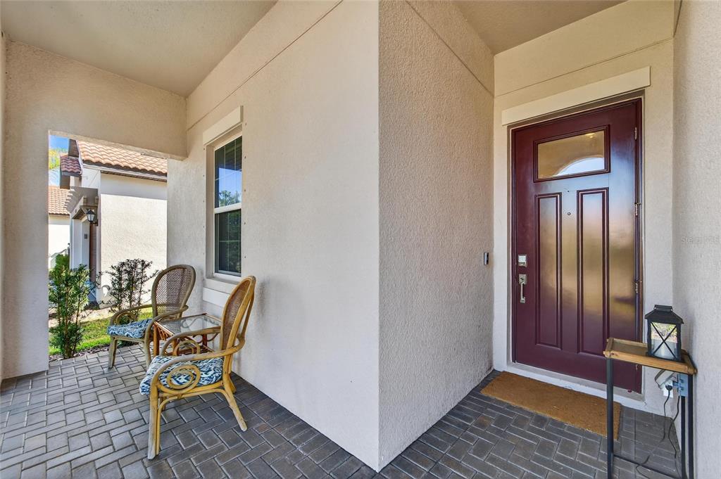 A covered front porch features a dark red door, brick pavers, and two wicker chairs — 17212 HOLLY WELL AVENUE, WIMAUMA, FL