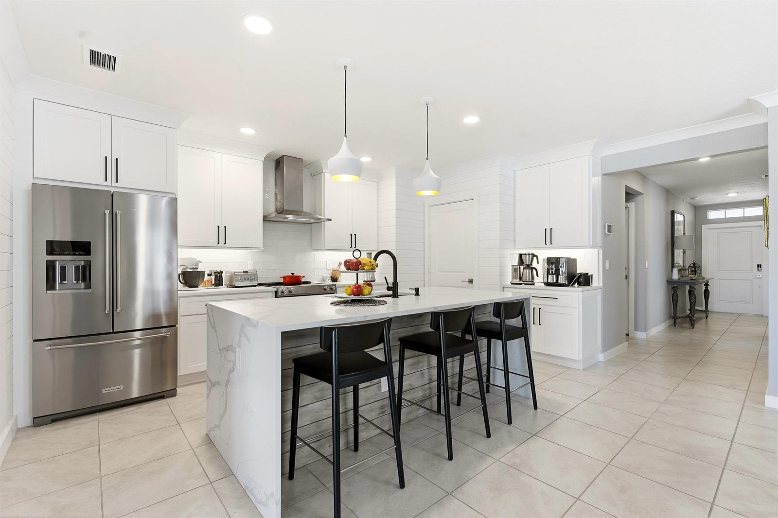 A modern white kitchen features a marble waterfall island, stainless steel appliances, and black stools — 3899 SE Lee Street, Stuart FL 34997, STUART, FL
