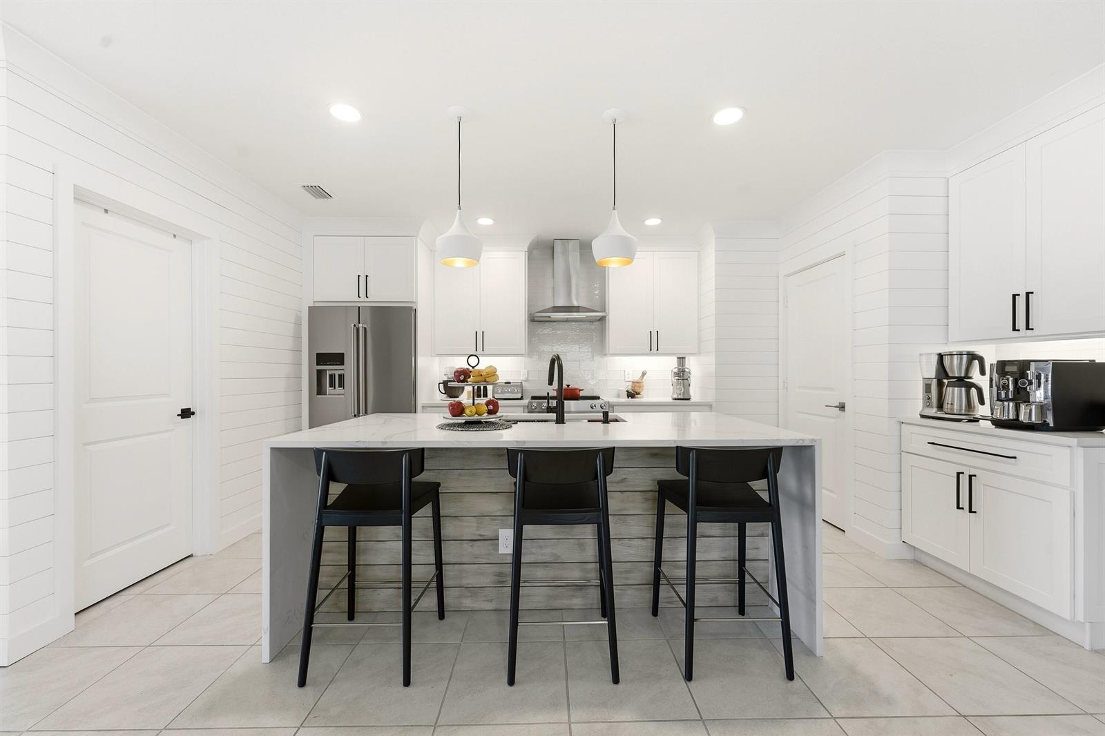 A modern white kitchen features a marble-topped island with three black stools and shiplap walls — 3899 SE Lee Street, Stuart FL 34997, STUART, FL