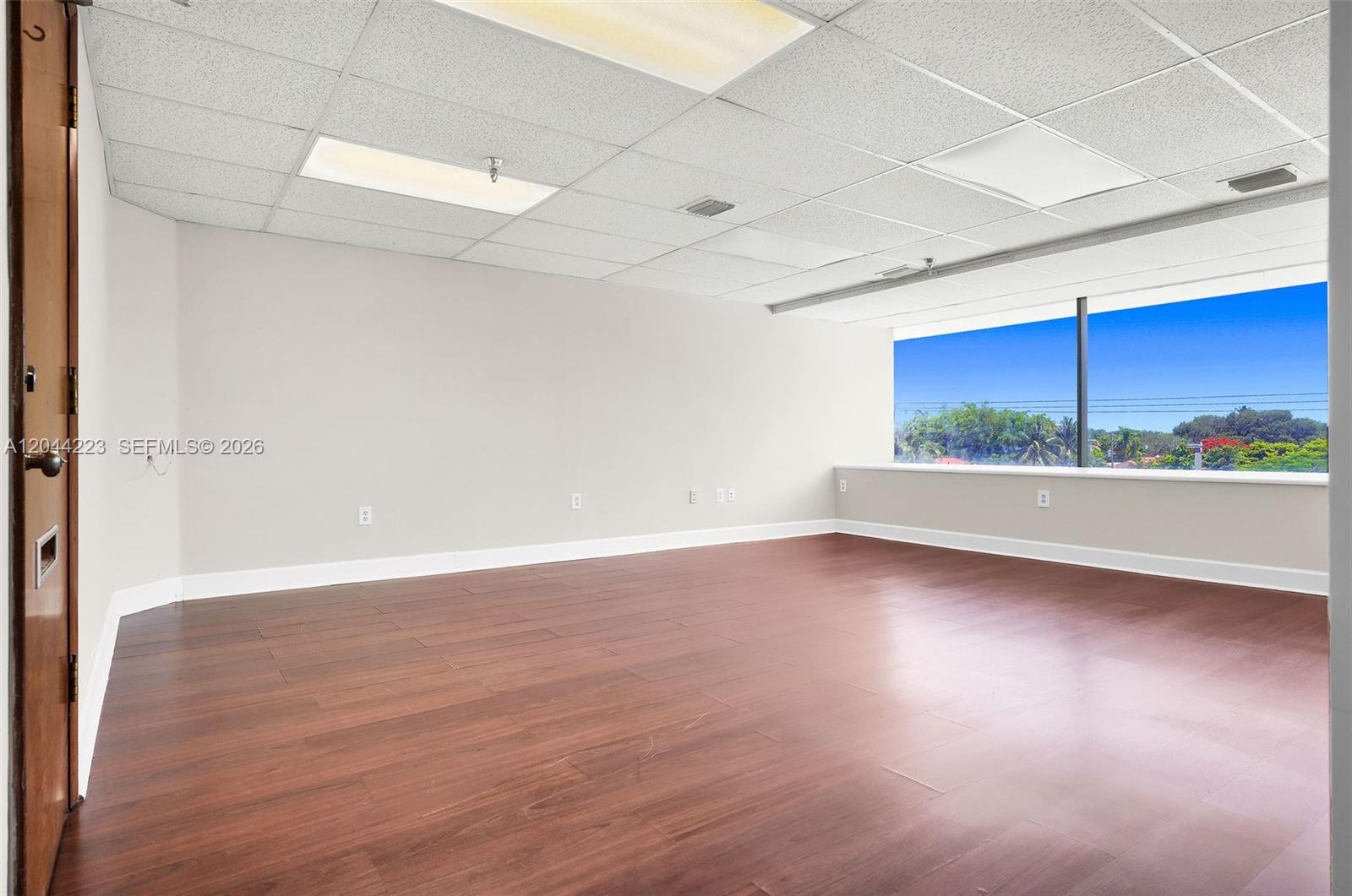 An empty office room features light gray walls, dark wood-look flooring, and a large window — 2150 SW 22nd St # 7A, Miami FL 33145, MIAMI, FL