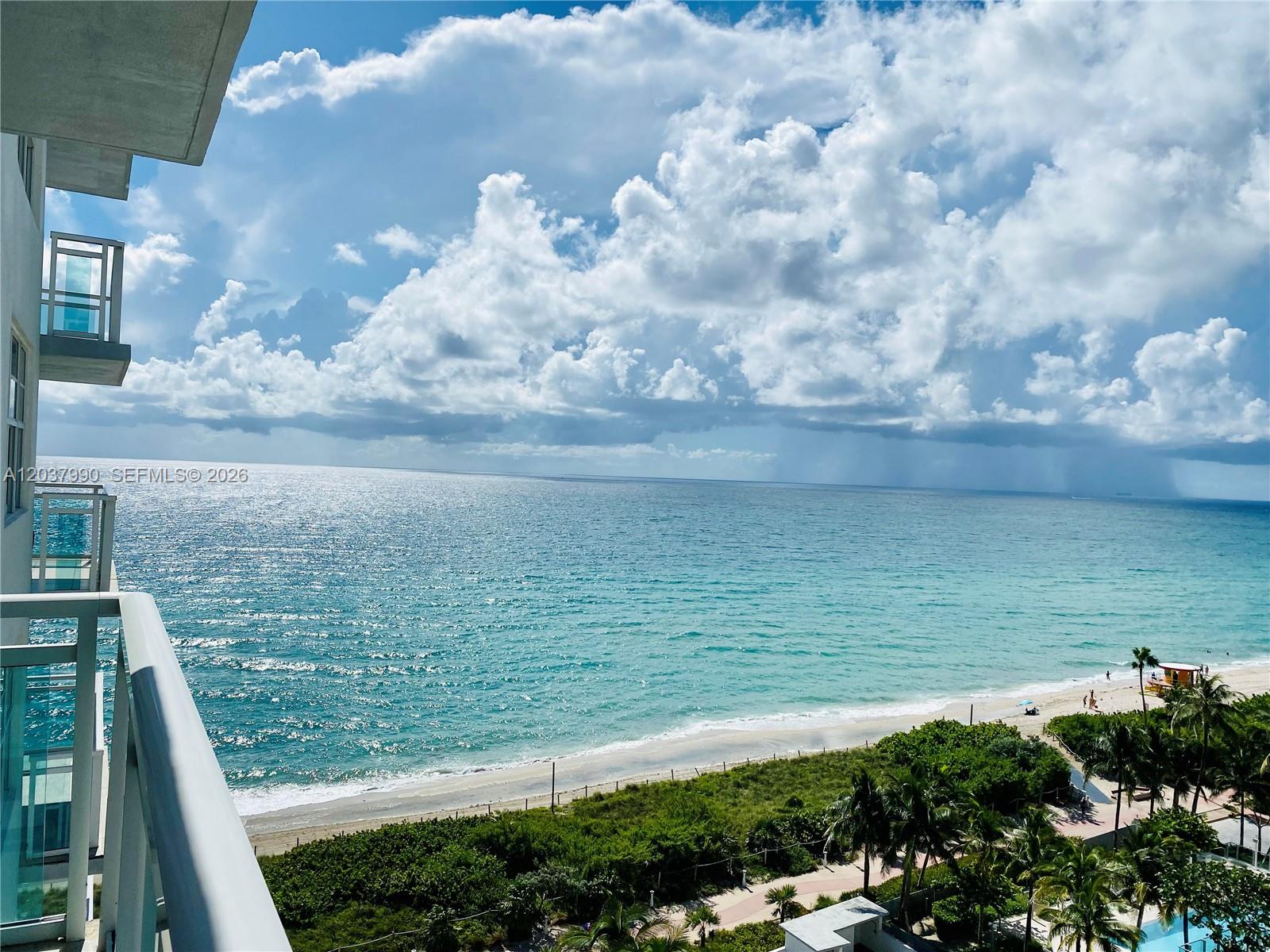 A high-angle view from a balcony overlooks a sandy beach, ocean, and cloudy sky — 6917 Collins Ave # 1205, Miami Beach FL 33141, MIAMI BEACH, FL