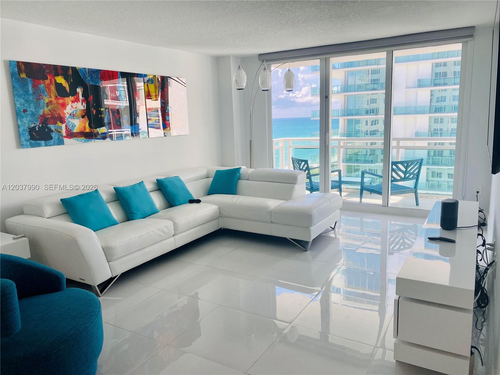 A white living room features a sectional sofa, blue accents, and a balcony overlooking the ocean — 6917 Collins Ave # 1205, Miami Beach FL 33141, MIAMI BEACH, FL