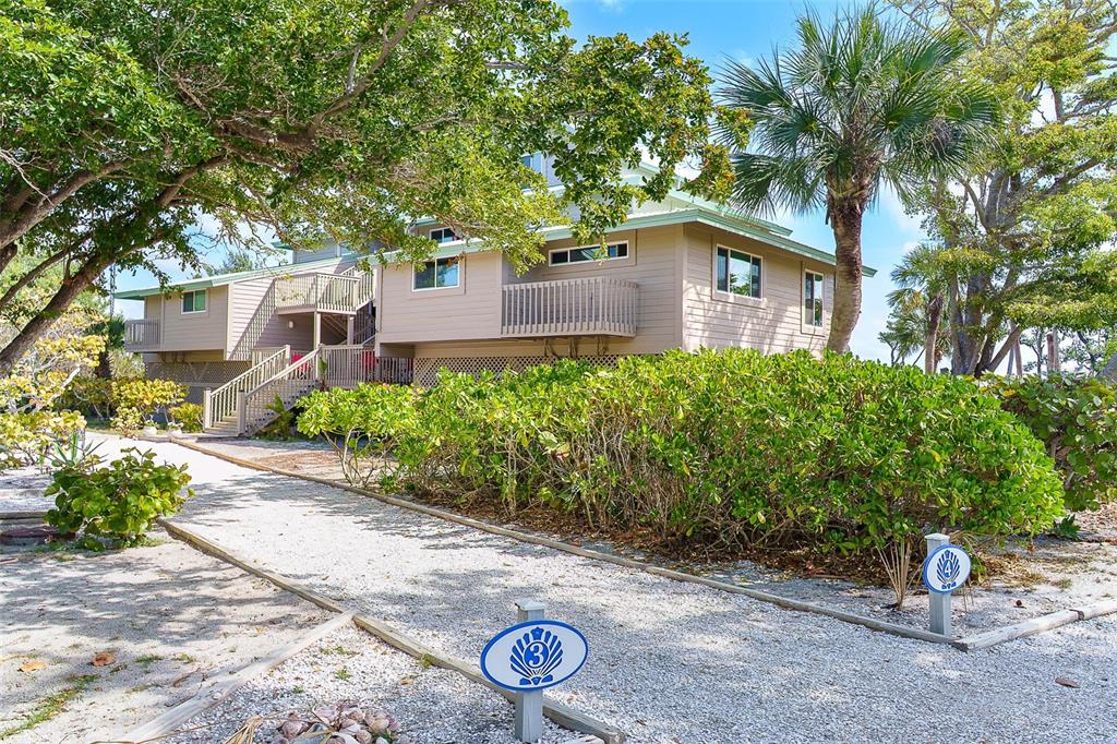 A two-story house with beige siding stands behind a gravel path and lush green bushes — 9200 LITTLE GASPARILLA ISLAND UNIT 403, PLACIDA, FL