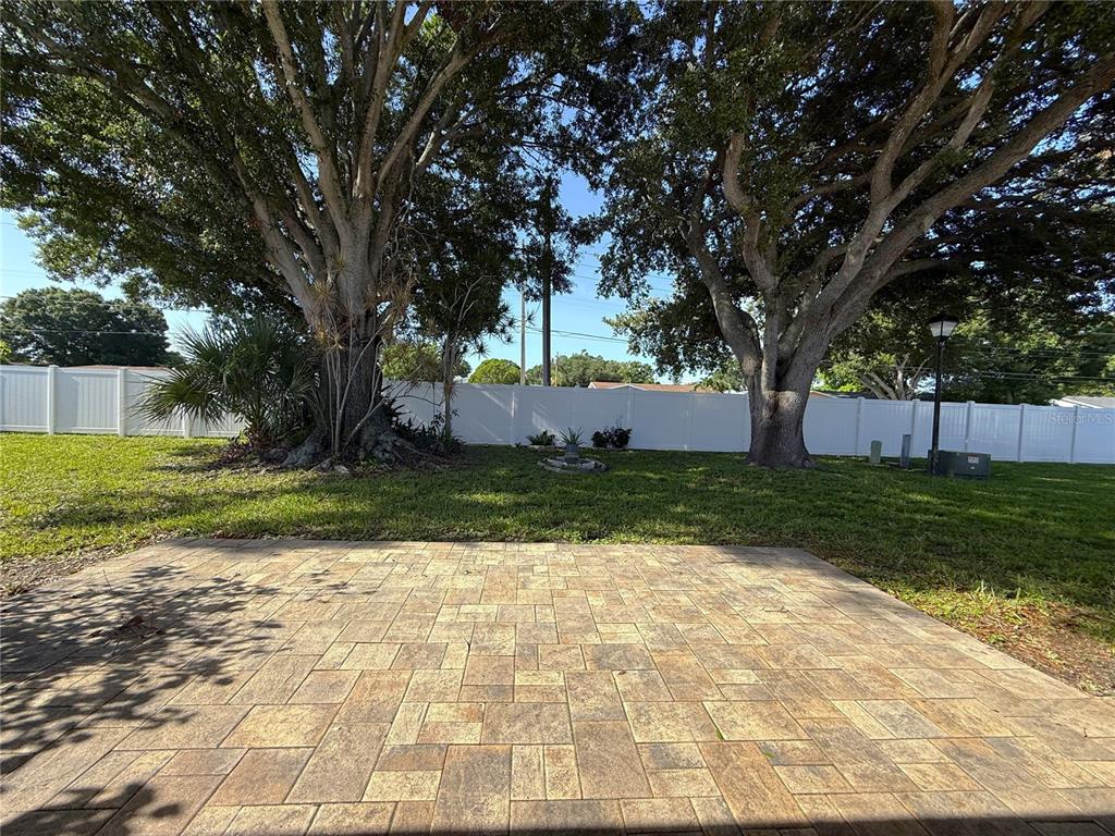 A paved patio overlooks a grassy backyard enclosed by a white vinyl fence and trees — 5420 PALM CREST COURT N, PINELLAS PARK, FL