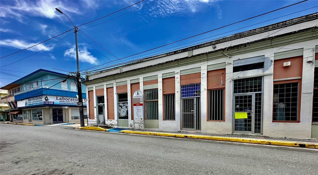 A street-level view of a commercial building with multiple storefronts and a blue laboratory building — 58 MUÑOZ RIVERA ESQ CORCHADO, JUNCOS, PR