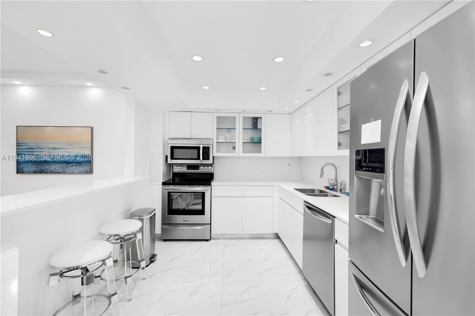 A modern white kitchen features stainless steel appliances, marble-look tile flooring, and a breakfast bar — 9455 Collins Ave # 806, Surfside FL 33154, SURFSIDE, FL