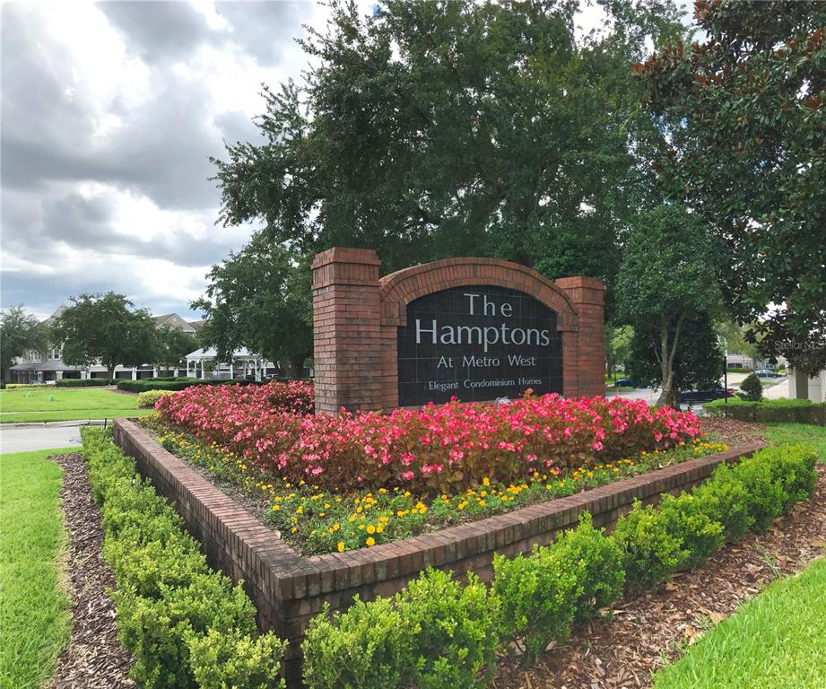 A brick monument sign for The Hamptons at Metro West sits amidst landscaped flower beds — 6627 QUEENS BOROUGH AVENUE UNIT 108, ORLANDO, FL
