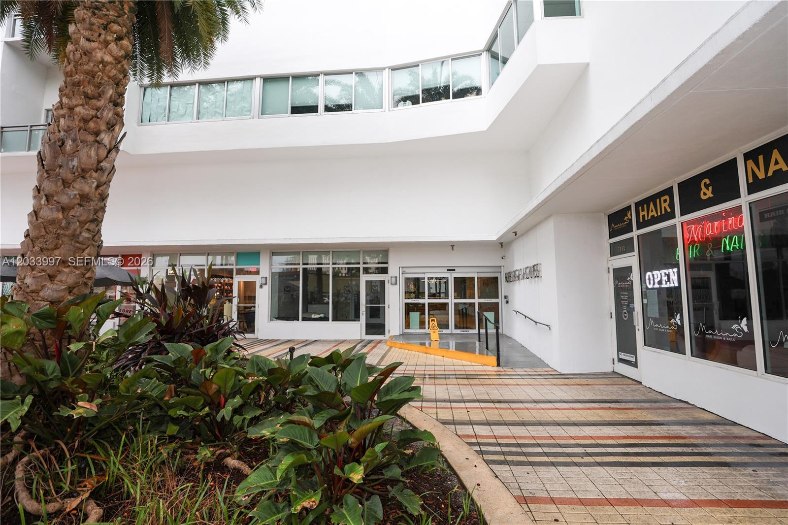 The exterior of a white commercial building features a palm tree and storefront windows — 7135 Collins Ave # 735, Miami Beach FL 33141, MIAMI BEACH, FL