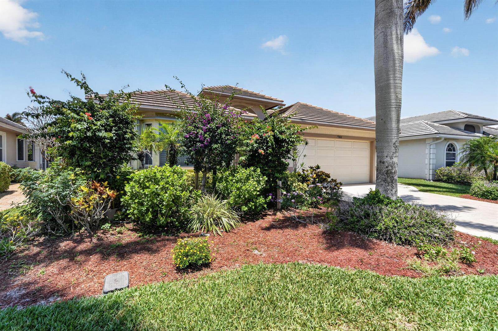 A single-story house with a beige exterior, a two-car garage, and lush front yard landscaping — 7982 SE Hempstead Circle, Hobe Sound FL 33455, HOBE SOUND, FL