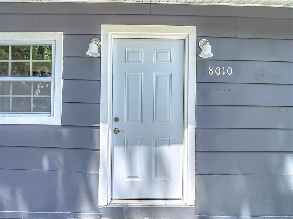 A white six-panel front door is centered on a gray horizontal-sided exterior wall — 8010 N 13TH STREET, TAMPA, FL