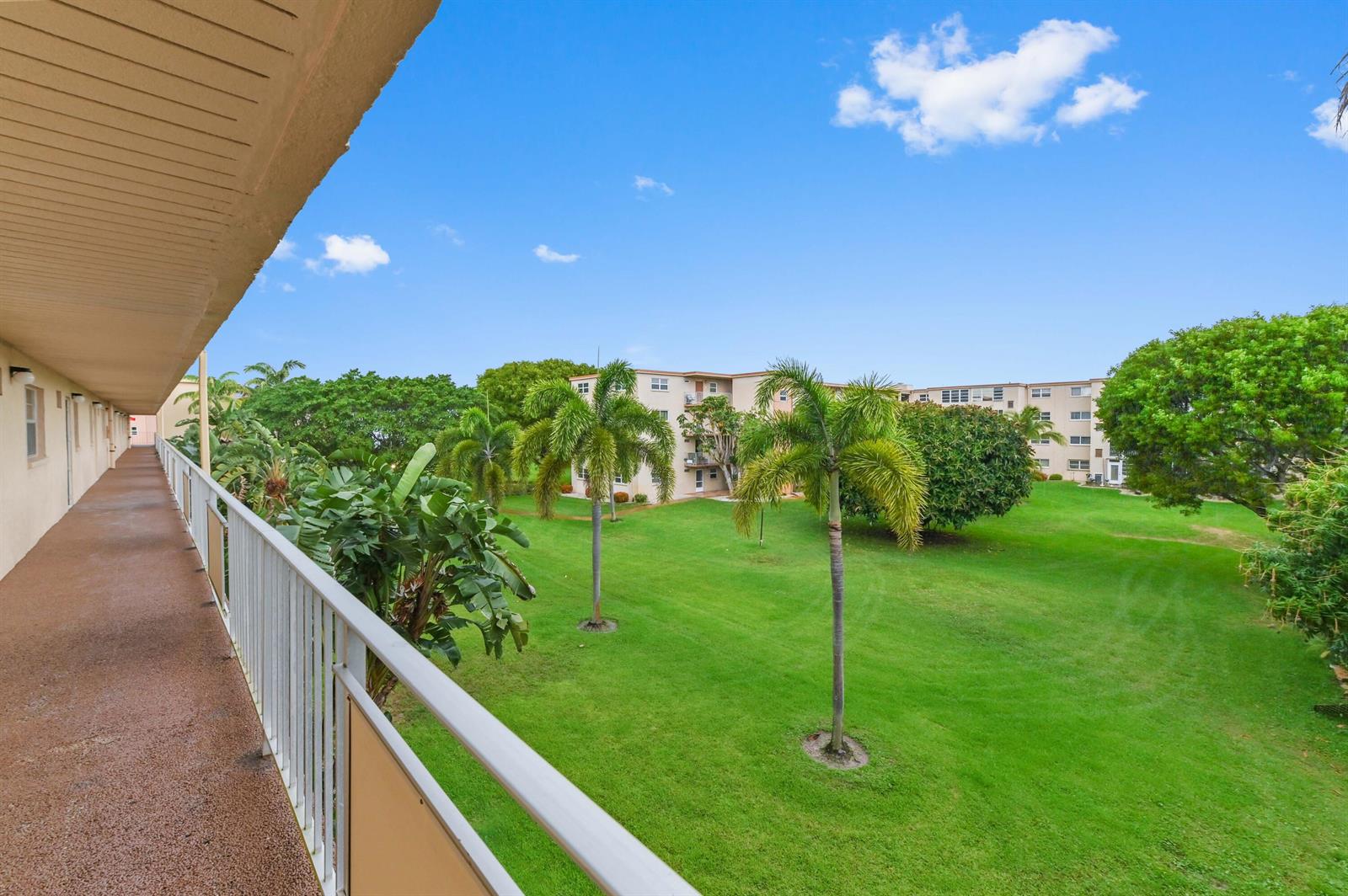 An elevated outdoor walkway overlooks a grassy courtyard with palm trees and apartment buildings — 2615 NE 3rd Court # 3170, Boynton Beach FL 33435, BOYNTON BEACH, FL
