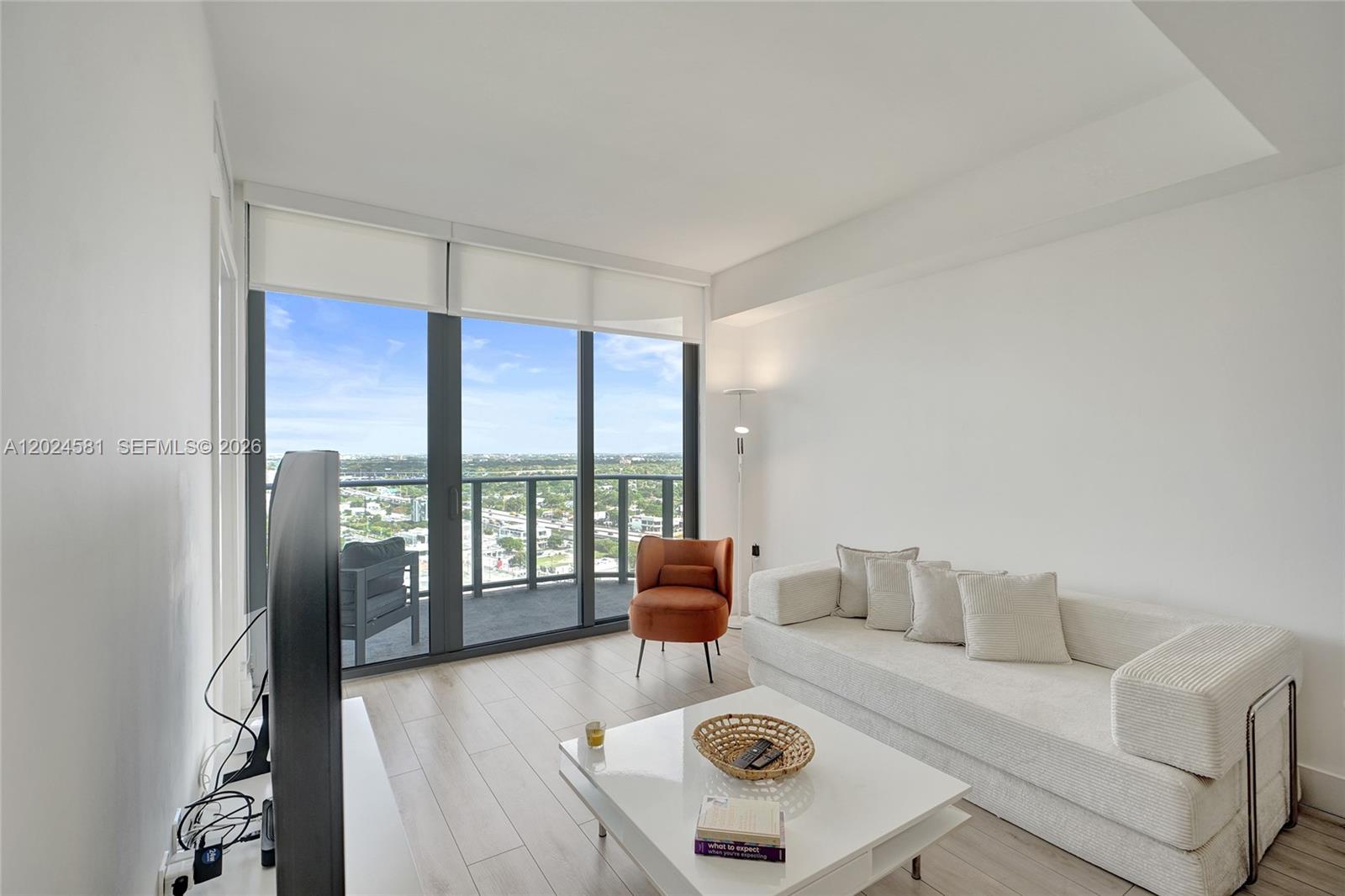 A modern living room features a white sofa, orange accent chair, and floor-to-ceiling balcony windows — 121 NE 34th St # 2310, Miami FL 33137, MIAMI, FL