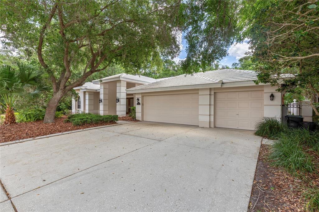 A beige single-story house features a three-car garage and a concrete driveway under large trees — 50 KELLEYS TRAIL, OLDSMAR, FL
