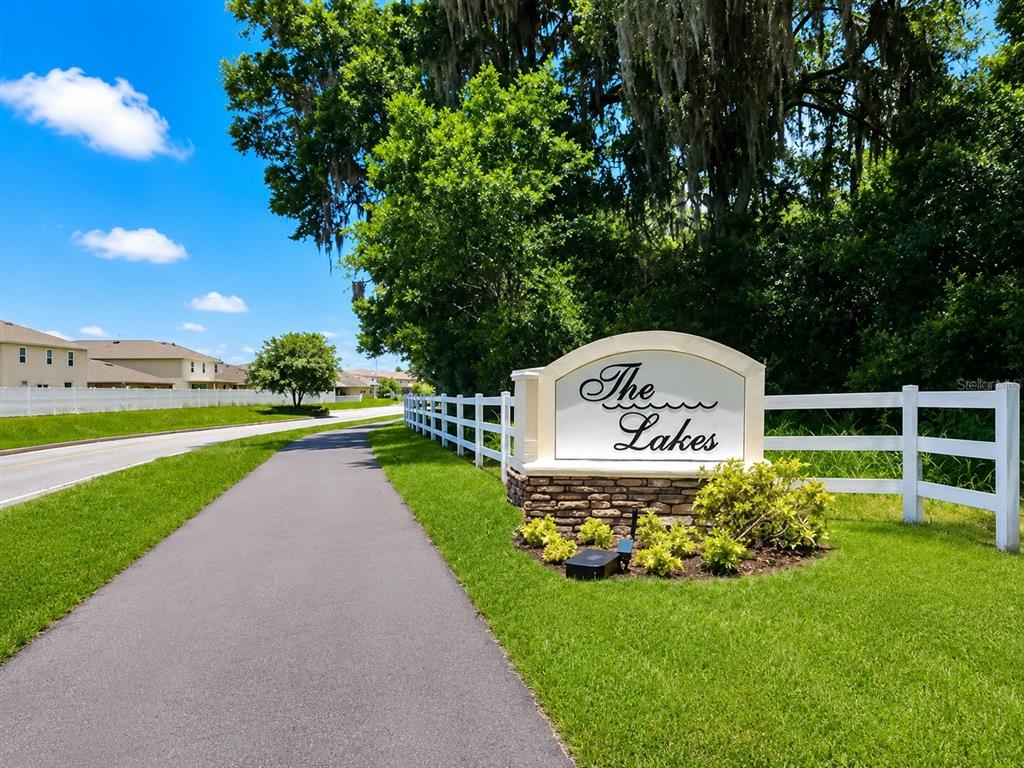 A paved path leads to a stone monument sign for The Lakes beside a white fence — 1002 PAISLEY STREET, LAKE ALFRED, FL