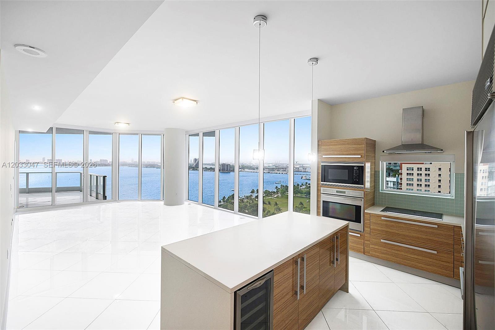 A modern kitchen with wood cabinetry and a white island overlooks a waterfront city view — 2020 N Bayshore Dr # 1602, Miami FL 33137, MIAMI, FL