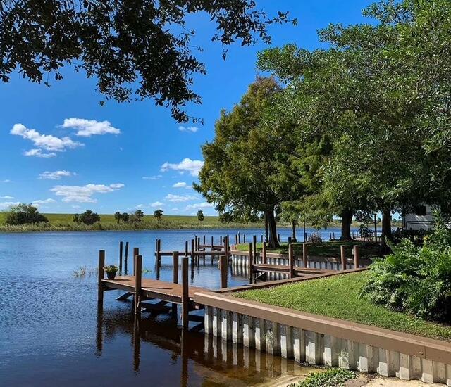 A wooden dock extends into a calm lake surrounded by trees and a grassy shoreline — 8680 Us-441, Okeechobee FL 34974, OKEECHOBEE, FL