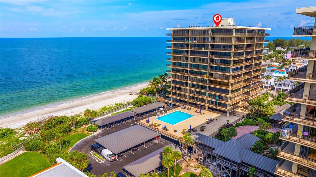 An aerial view shows a multi-story beachfront condominium building with a pool and parking area — 7650 BAYSHORE DRIVE UNIT 1005, TREASURE ISLAND, FL