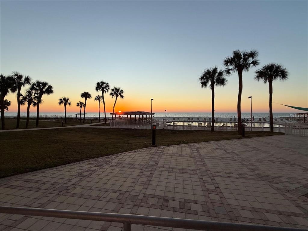 A paved patio overlooks a swimming pool, palm trees, and the ocean at sunset — 880 MANDALAY AVENUE UNIT C410, CLEARWATER, FL