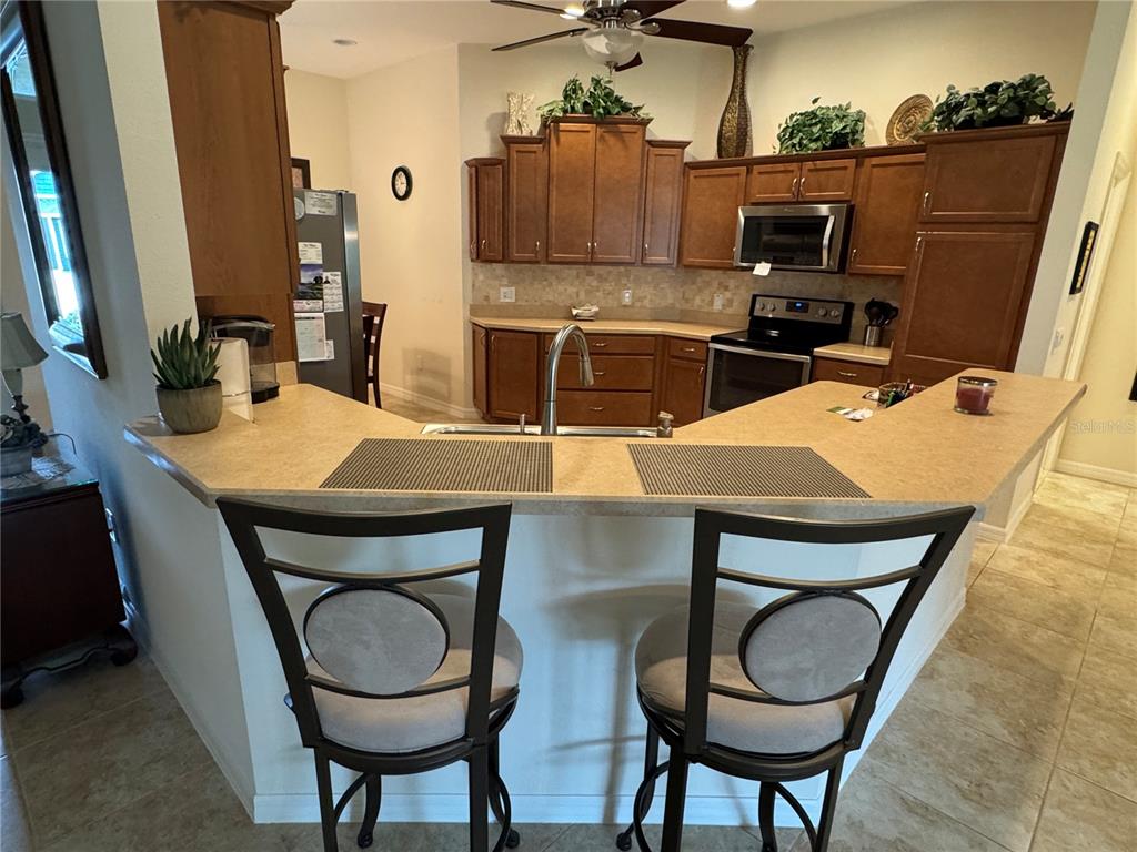 A kitchen features brown cabinets, tan countertops, a breakfast bar with two stools, and appliances — 939 KOLLER COURT, THE VILLAGES, FL