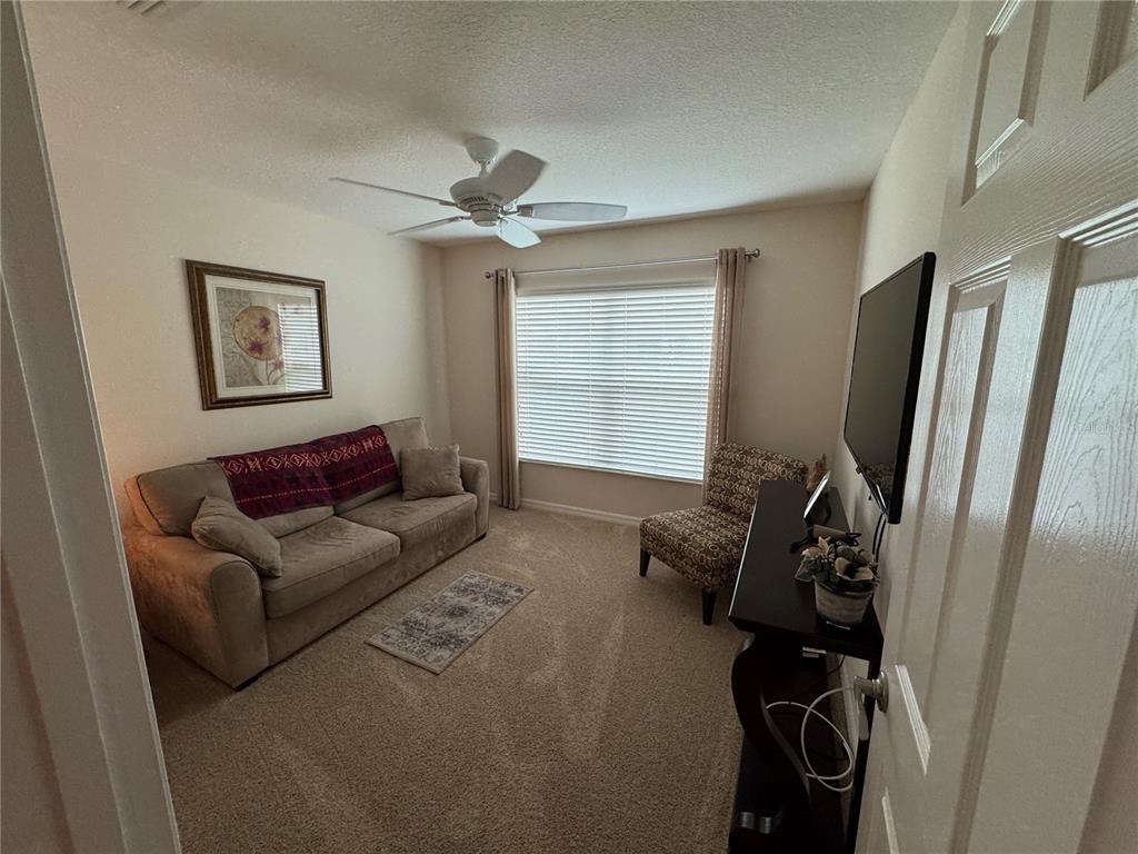 A carpeted living room features a sofa, a patterned armchair, a television, and a ceiling fan — 939 KOLLER COURT, THE VILLAGES, FL