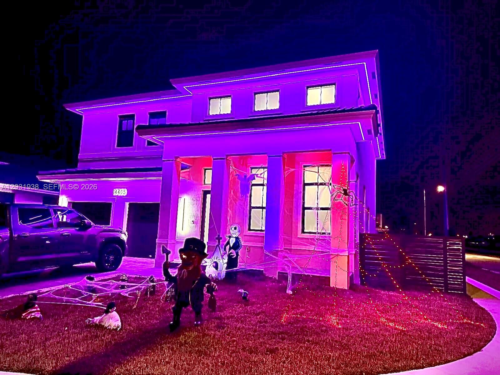 A two-story house exterior is decorated with purple lighting and Halloween-themed lawn ornaments at night — 14405 SW 13th Ter, Miami FL 33184, MIAMI, FL