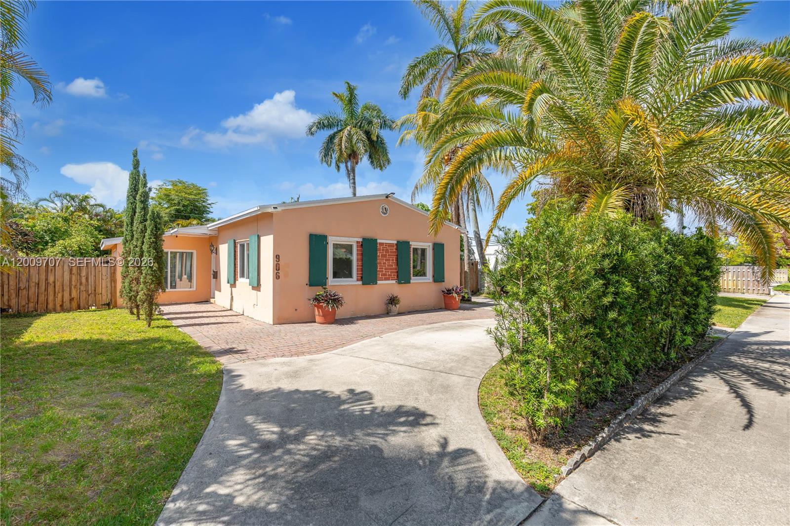 A single-story peach-colored house features a paved driveway, green shutters, and surrounding palm trees — 906 NE 5th St, Hallandale Beach FL 33009, HALLANDALE BEACH, FL