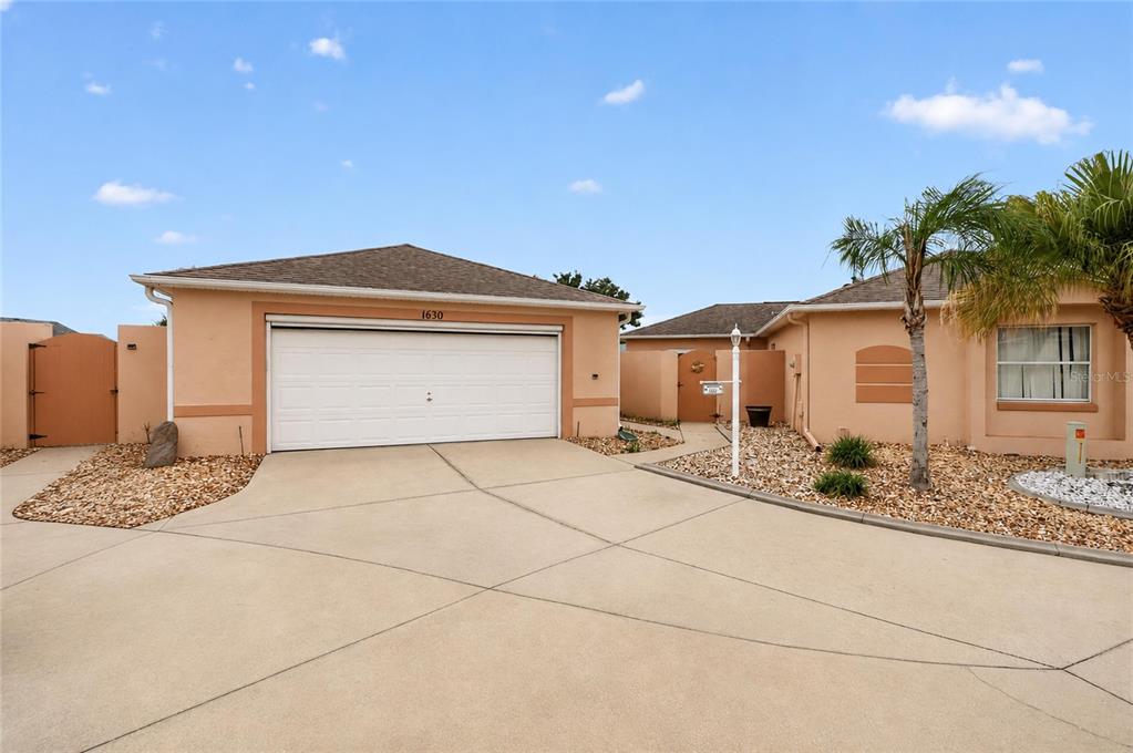 A single-story house with a white garage door, tan stucco walls, and a gravel yard — 1630 TAMPICO PLACE, LADY LAKE, FL