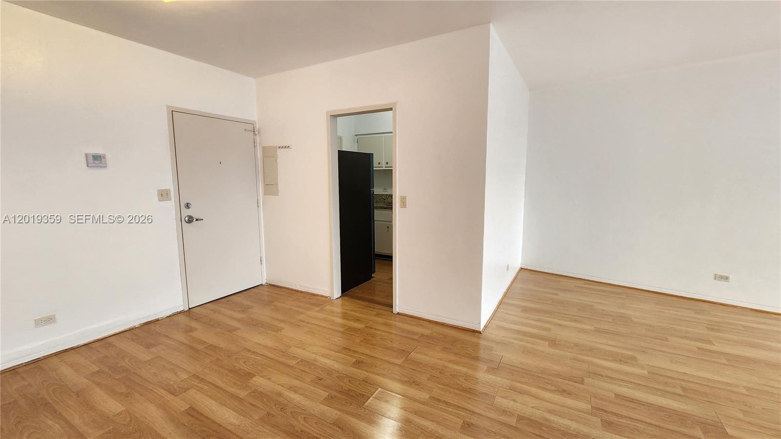 An empty room with light wood-look flooring, white walls, and a doorway leading to a kitchen — 7441 Wayne # O, Miami Beach FL 33141, MIAMI BEACH, FL