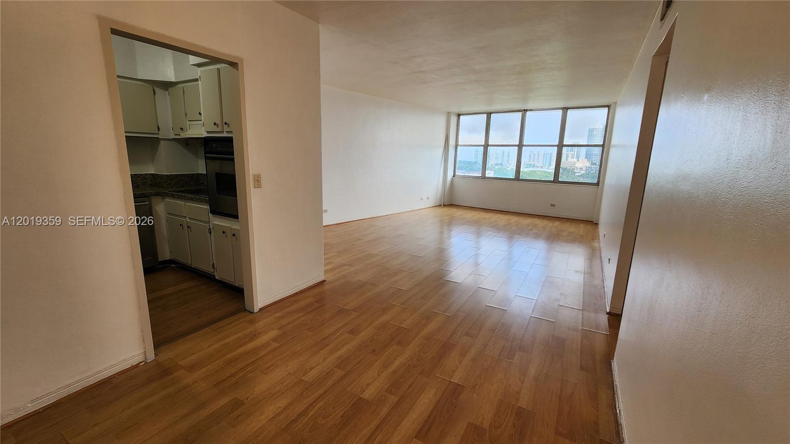 An empty living area with wood-look flooring leads to a kitchen and large windows — 7441 Wayne # O, Miami Beach FL 33141, MIAMI BEACH, FL